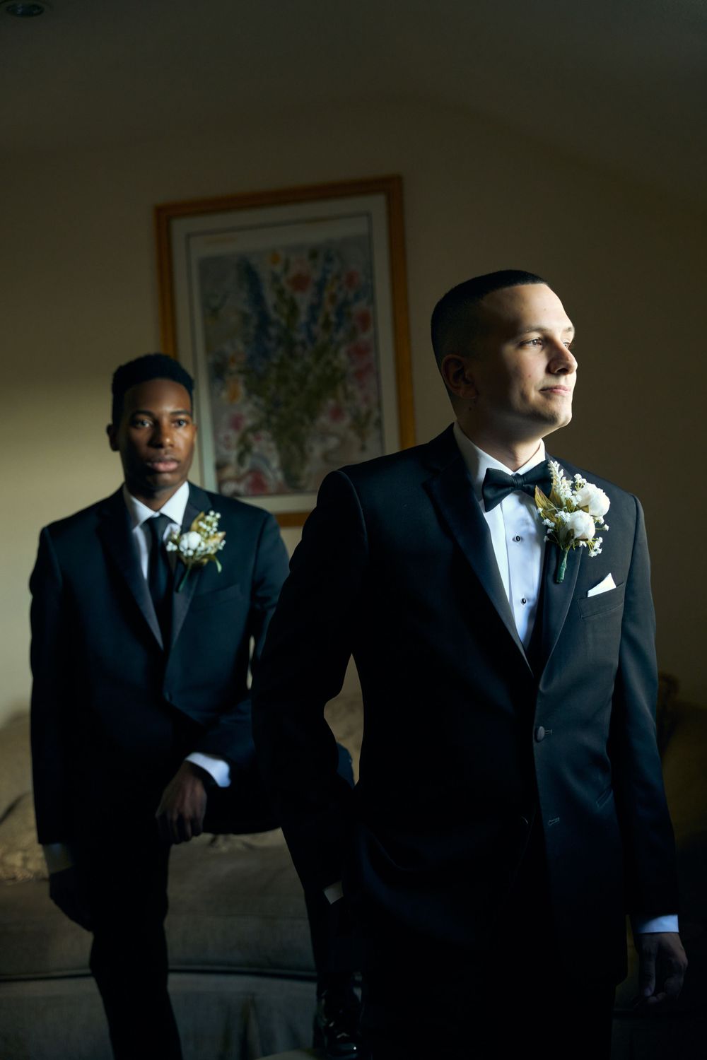Groomsmen in black suits and white boutonnieres stand in dramatic lighting against a wall with artwork.