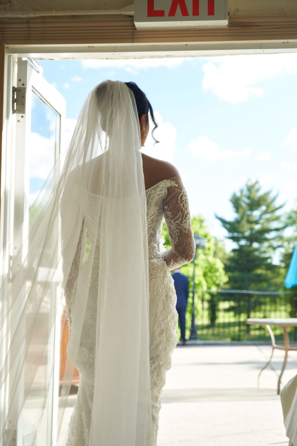 Row of flowing white wedding dresses and veils backlit by natural window light with an exit sign visible.