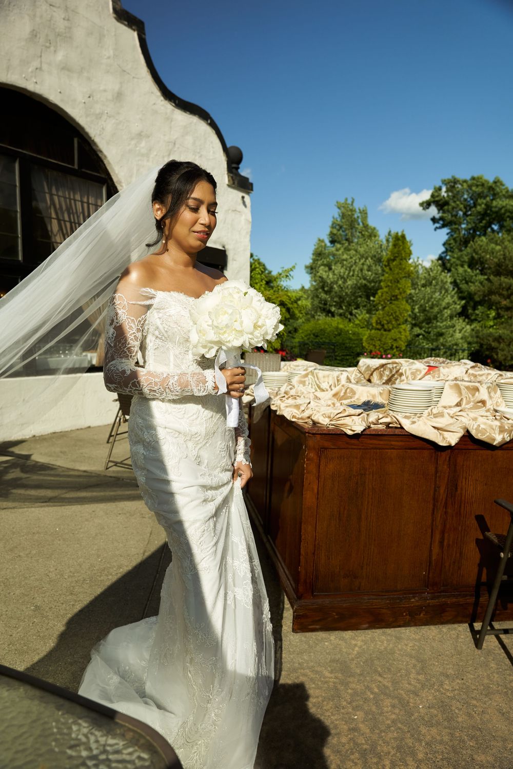 White lace wedding dress with flowing train displayed outdoors against rustic wooden backdrop.