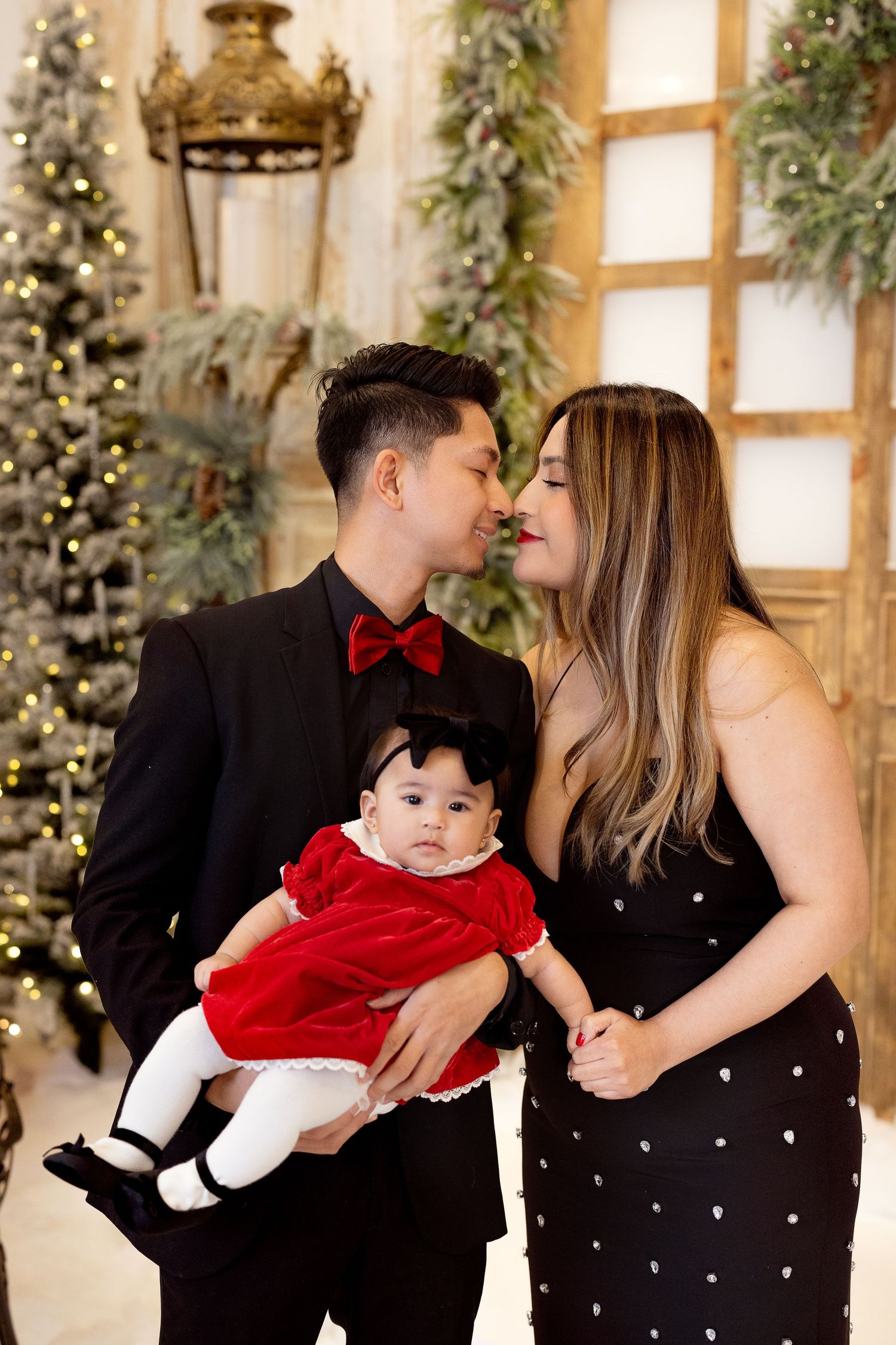 Elegant family photo with Christmas decor, featuring a baby in red dress, against a backdrop of trees and holiday lights.
