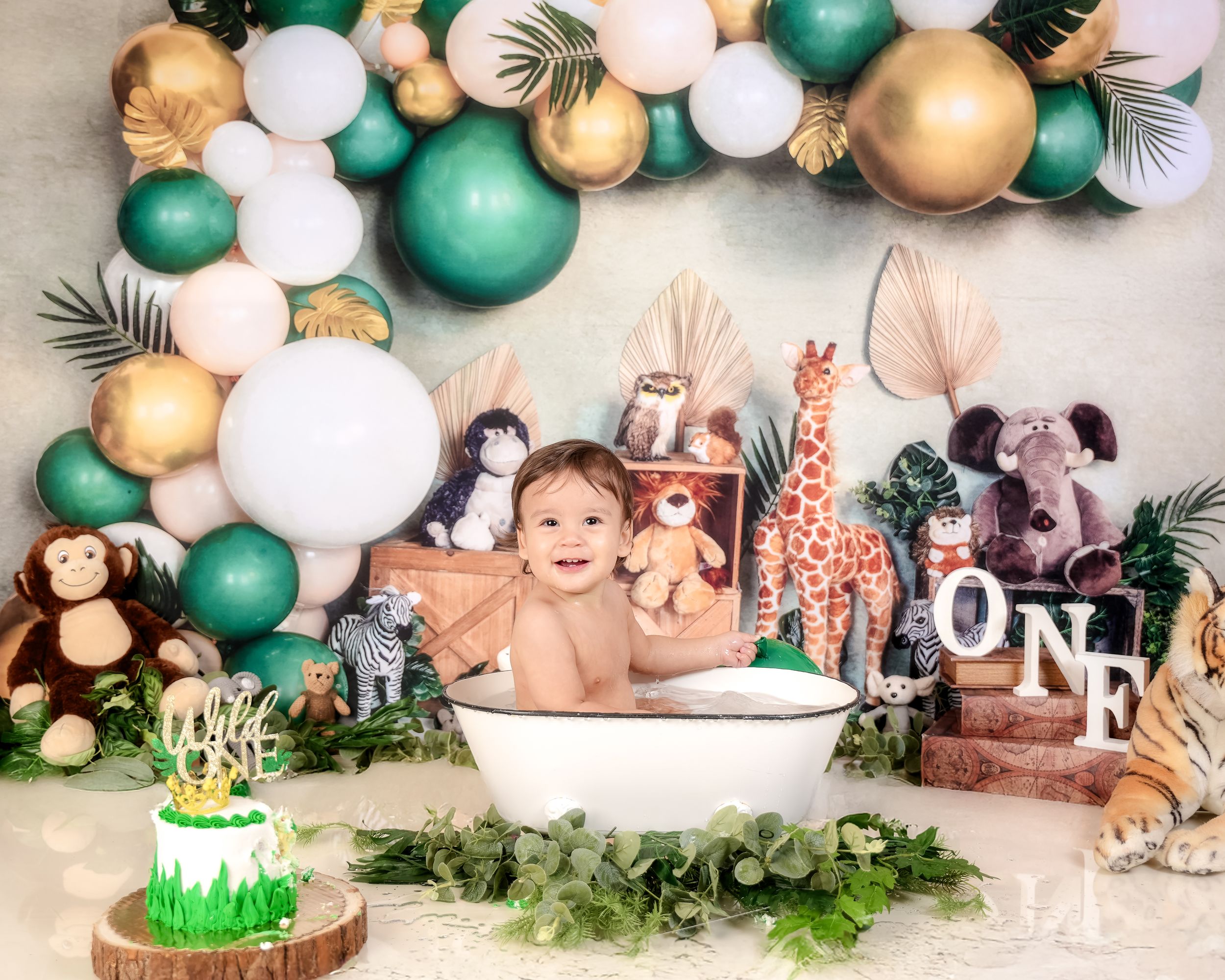 Safari-themed cake smash setup with teal and gold balloon arch, stuffed animals, and bathtub decorated with greenery.