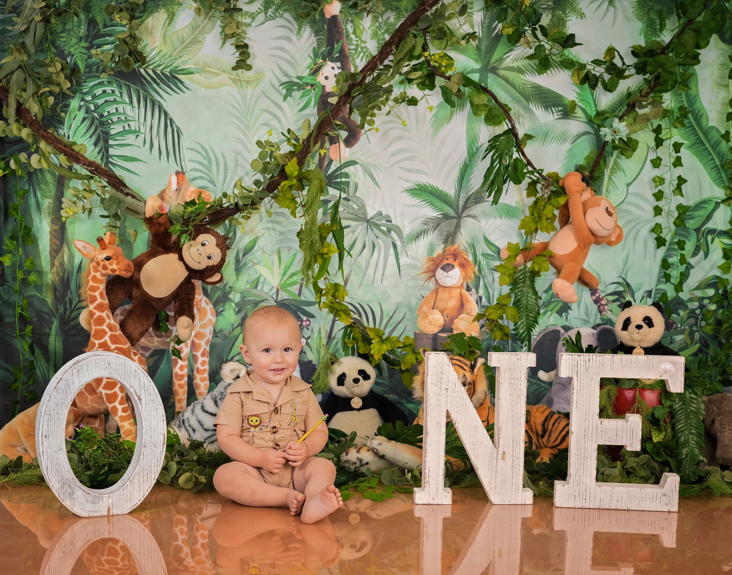 Baby sits by ONE letters with stuffed animals in jungle-themed first birthday photo shoot with lush greenery backdrop.