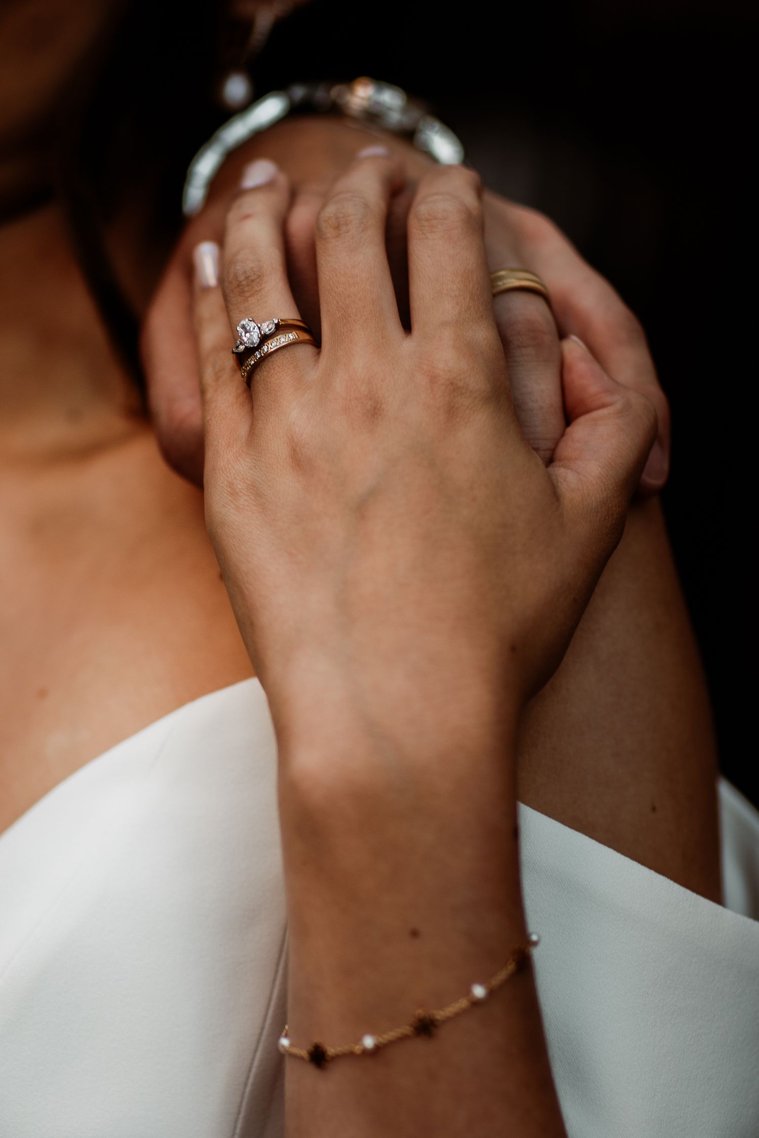 Close-up of hands wearing an engagement ring and delicate pearl bracelet against white wedding dress fabric.