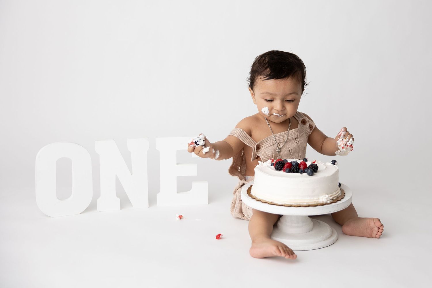Baby in diaper sits beside white cake with berries on cake stand during first birthday photoshoot against white backdrop.