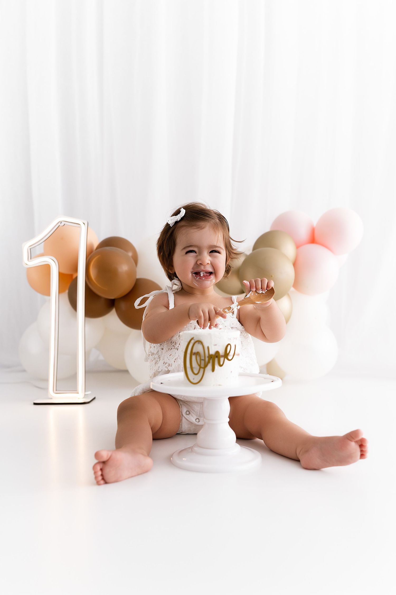Baby in white outfit sits by number one decoration with pink and gold balloons.