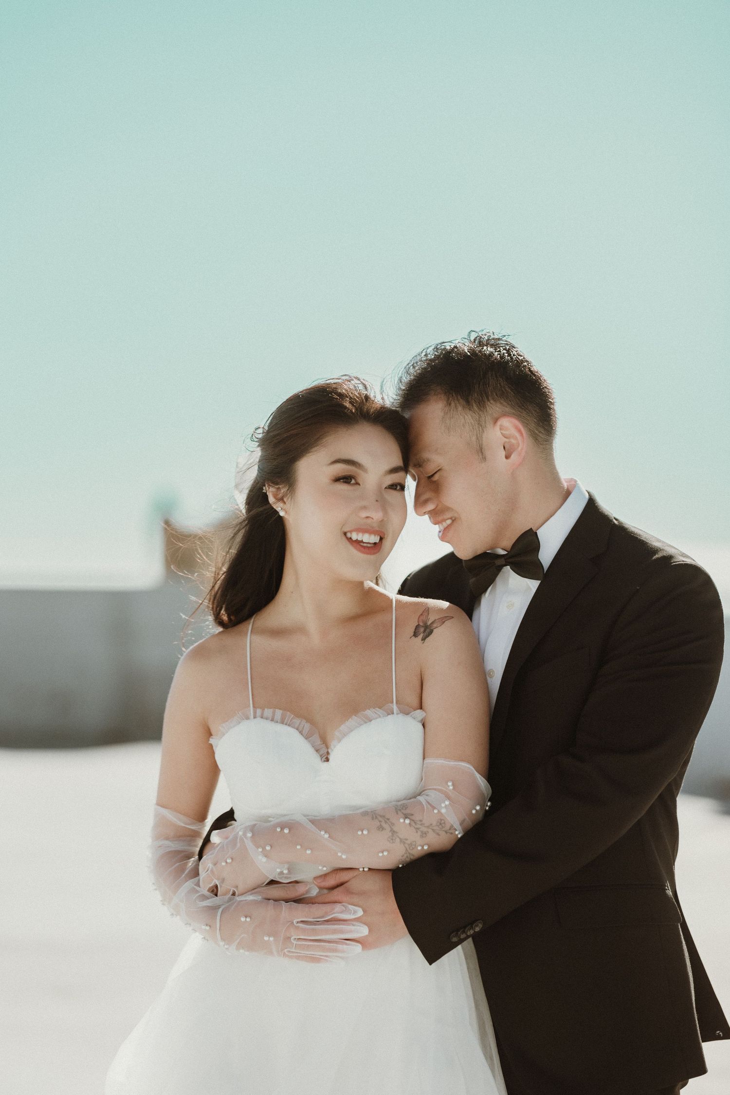 A romantic wedding photo with a couple embracing outdoors in formal attire against a light blue sky.