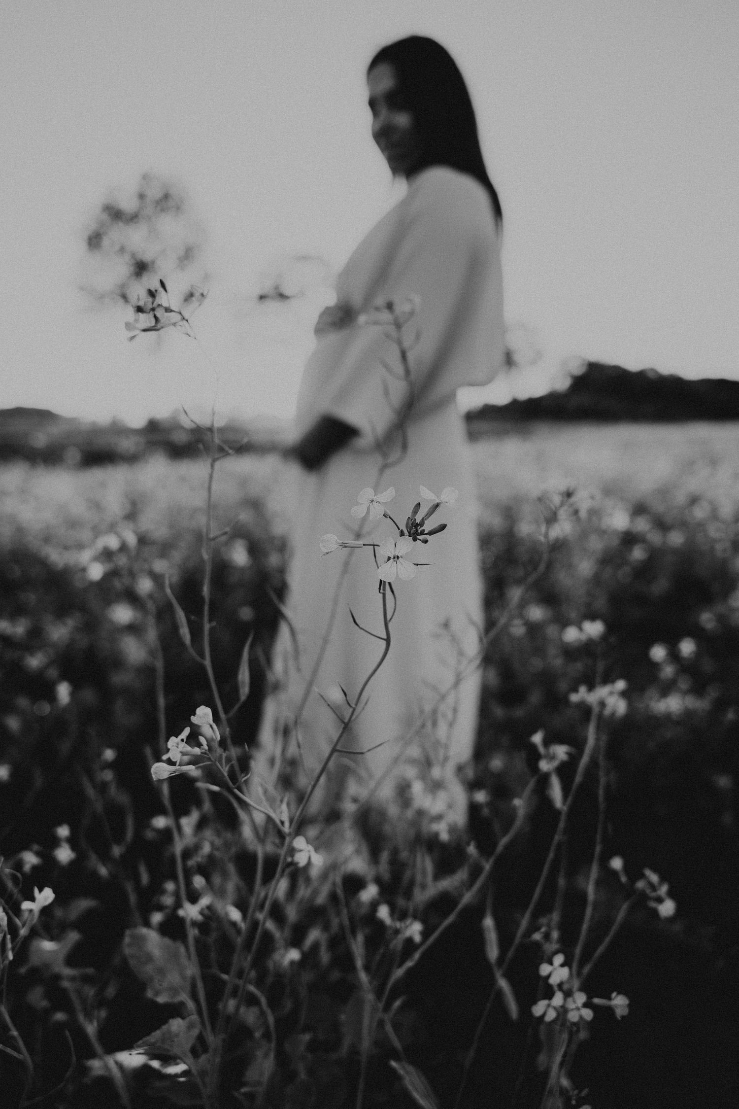 Zwart wit foto van een zwangere vrouw in een veld met bloemen, focus ligt op bloemen, vrouw is blurry zichtbaar.