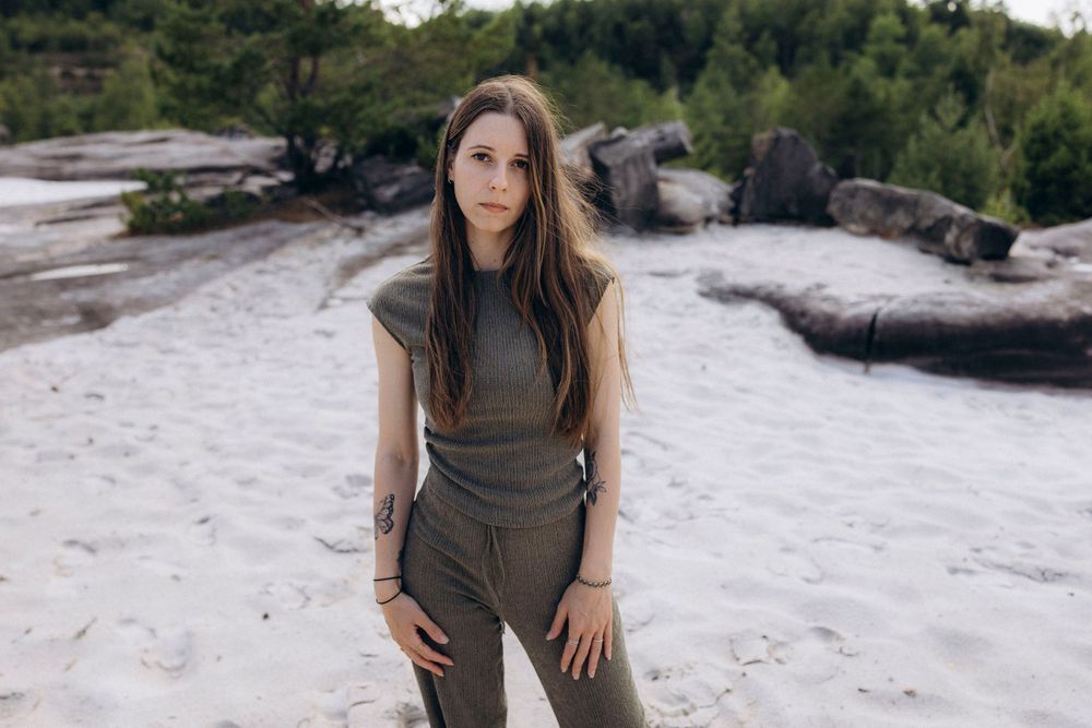 A person in a sleeveless grey outfit stands on a rocky beach with trees in the background.