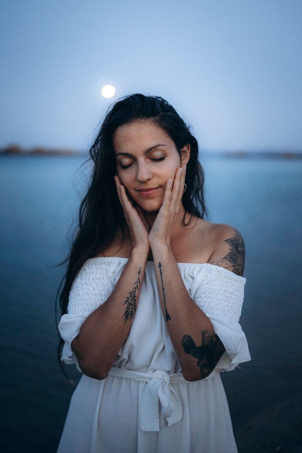 Woman in white off-shoulder dress with tattoos, hands on cheeks, eyes closed, standing by water under a full moon.