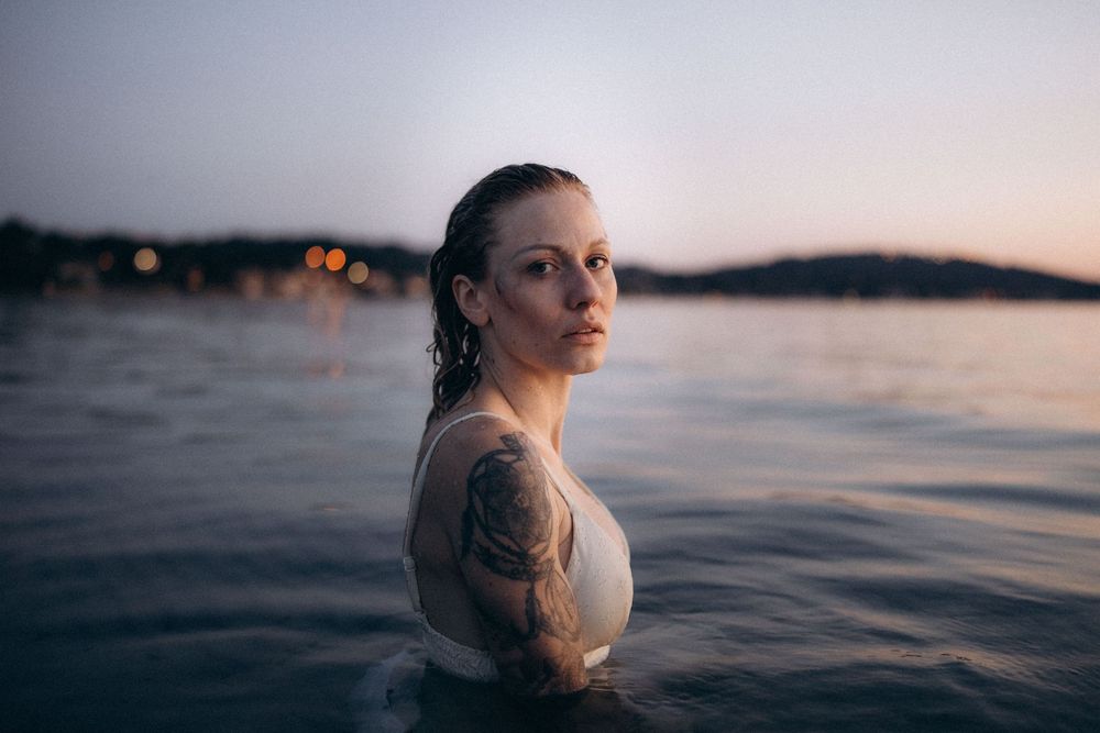 A person stands waist-deep in calm water at dusk with a moody sunset and distant shoreline in the background.
