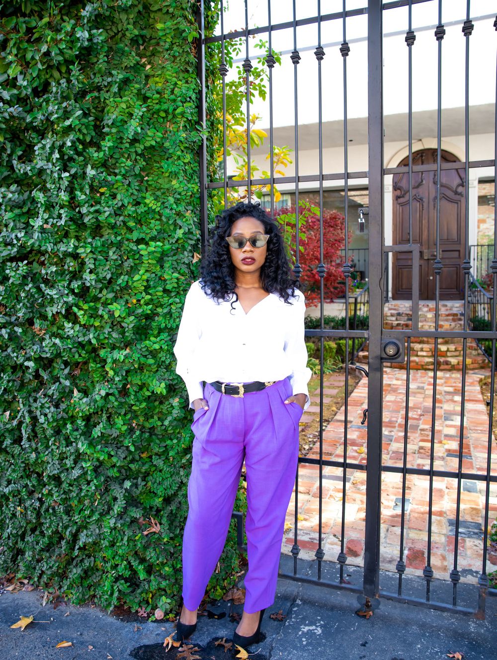 A person wearing a white sweater and purple pants poses in front of an ornate iron gate and climbing vines.