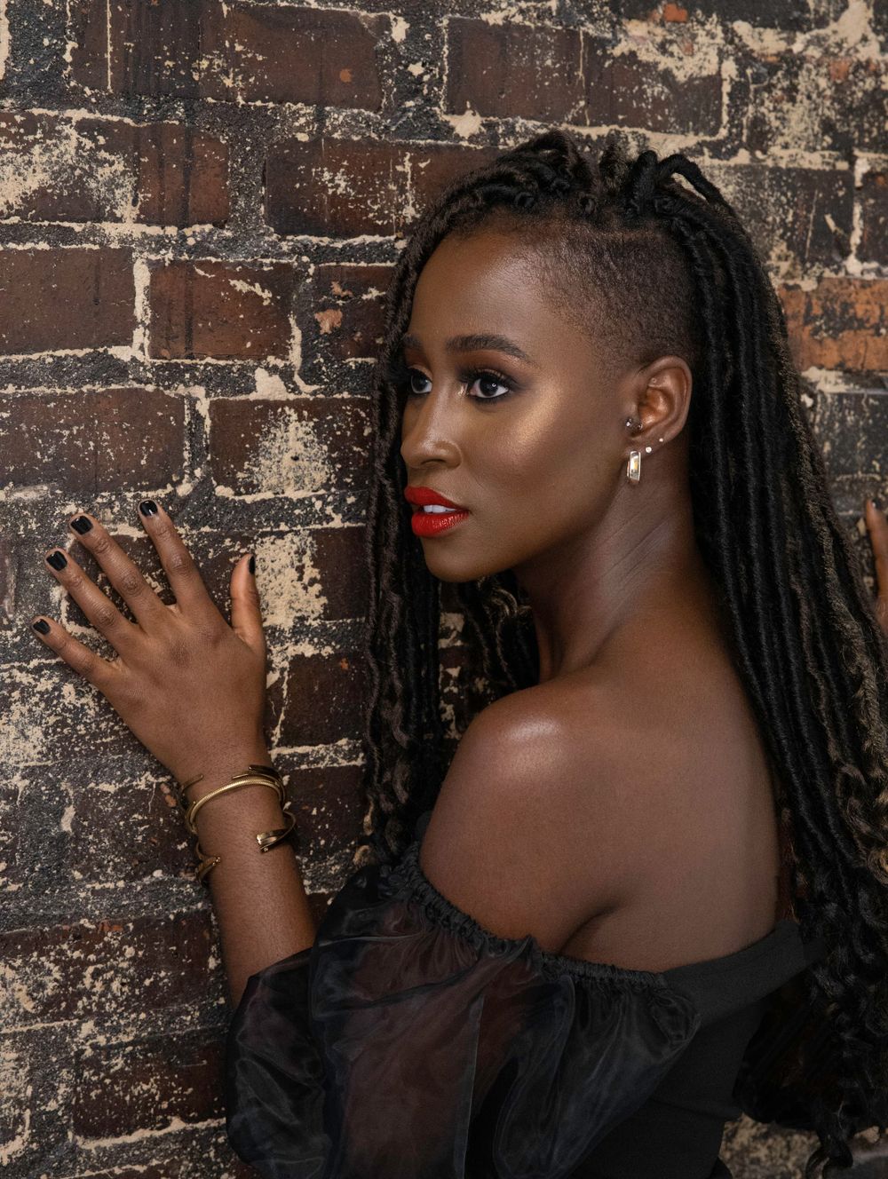 A portrait against a rustic brick wall featuring long braids, red lipstick and an elegant off-shoulder black top.