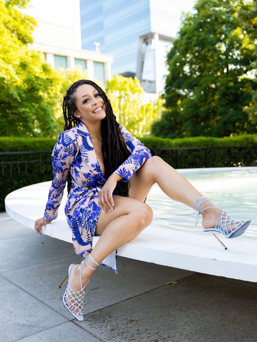 Stylish person in blue patterned dress and silver heels poses outdoors near modern building and greenery.