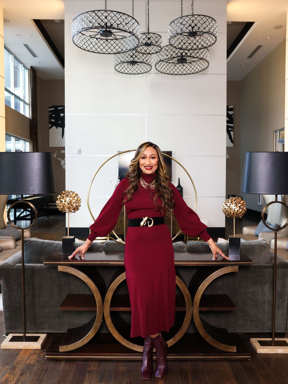 A person in a burgundy long-sleeved dress poses in a modern interior with circular light fixtures and gold accents.