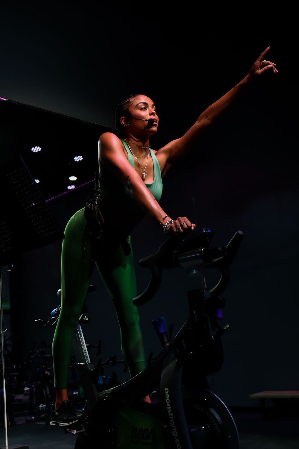 Exercise instructor leads an energetic spin class in a dark studio with dramatic lighting and raised arm gesture.