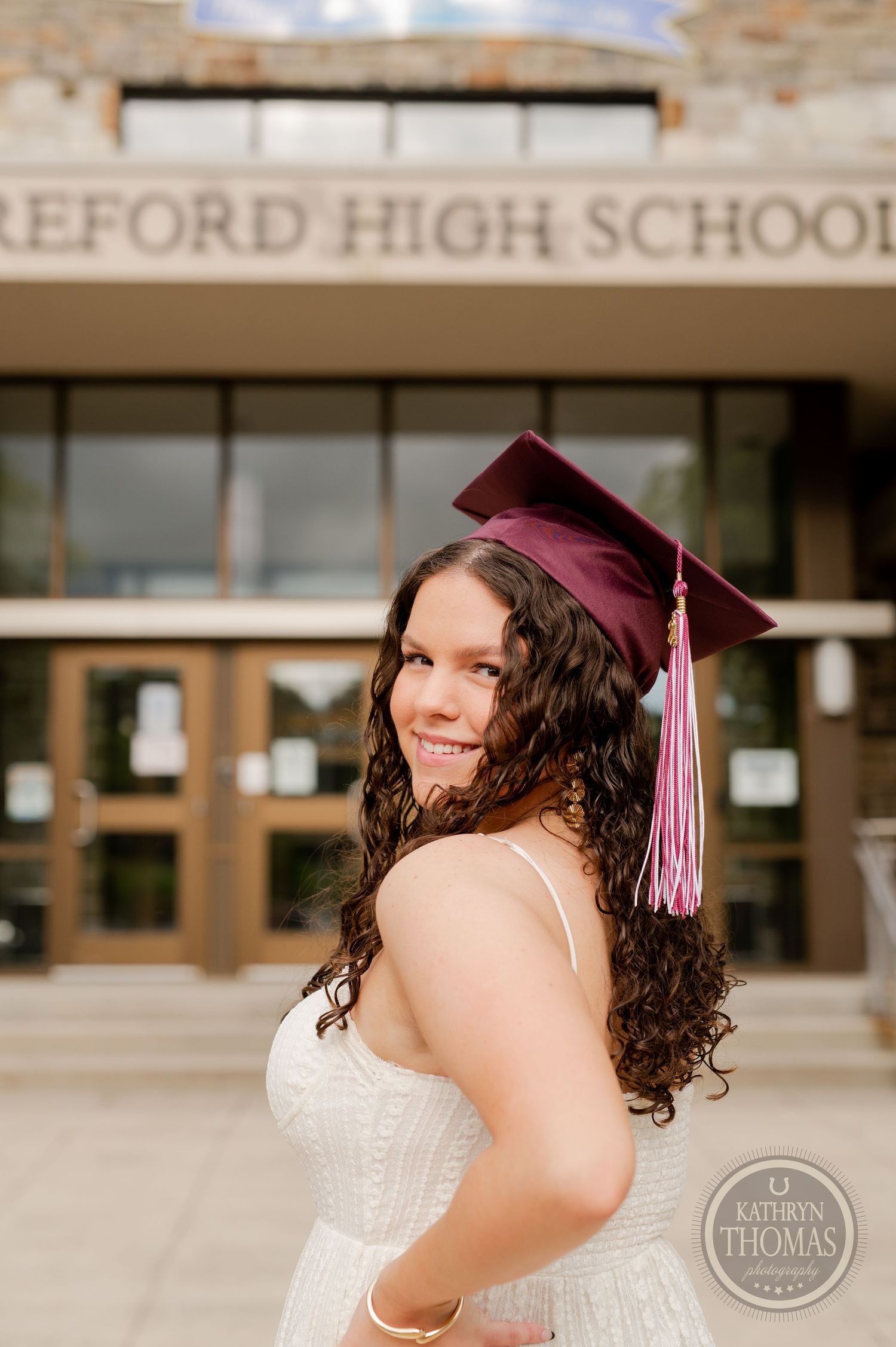 Graduate with curly hair smiles in white dress and maroon cap at school entrance.