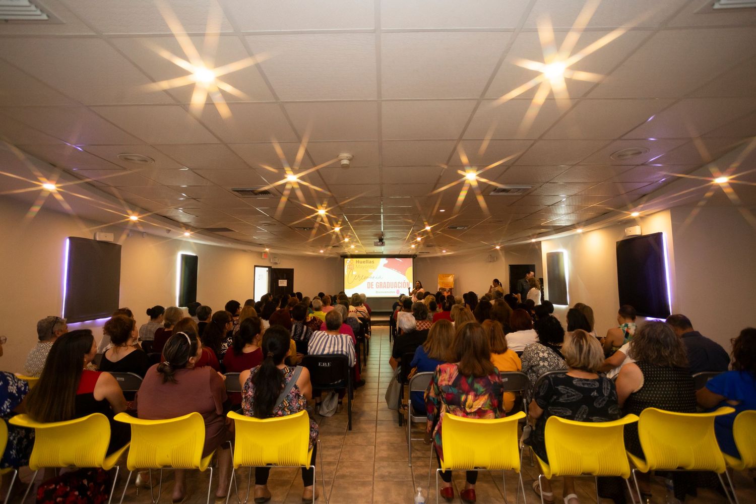 Conference attendees seated in yellow chairs watch a presentation in a room with decorative starburst ceiling lights.