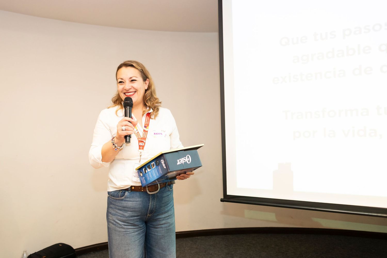 A presenter holds a microphone and folder while speaking in front of a projection screen in a meeting room.