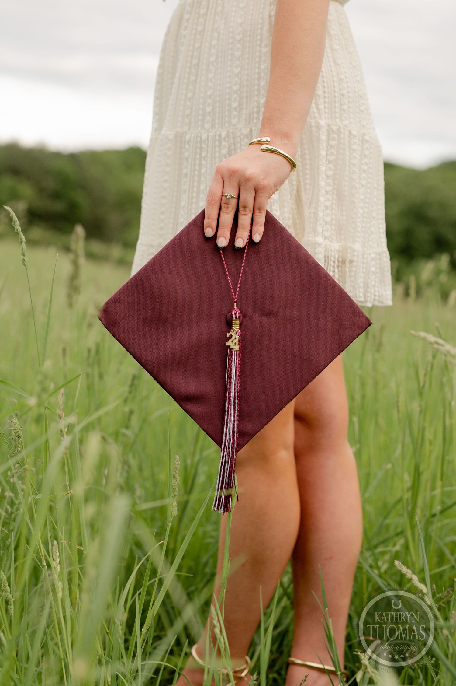 Hand holding maroon graduation cap with tassel against backdrop of tall grass.
