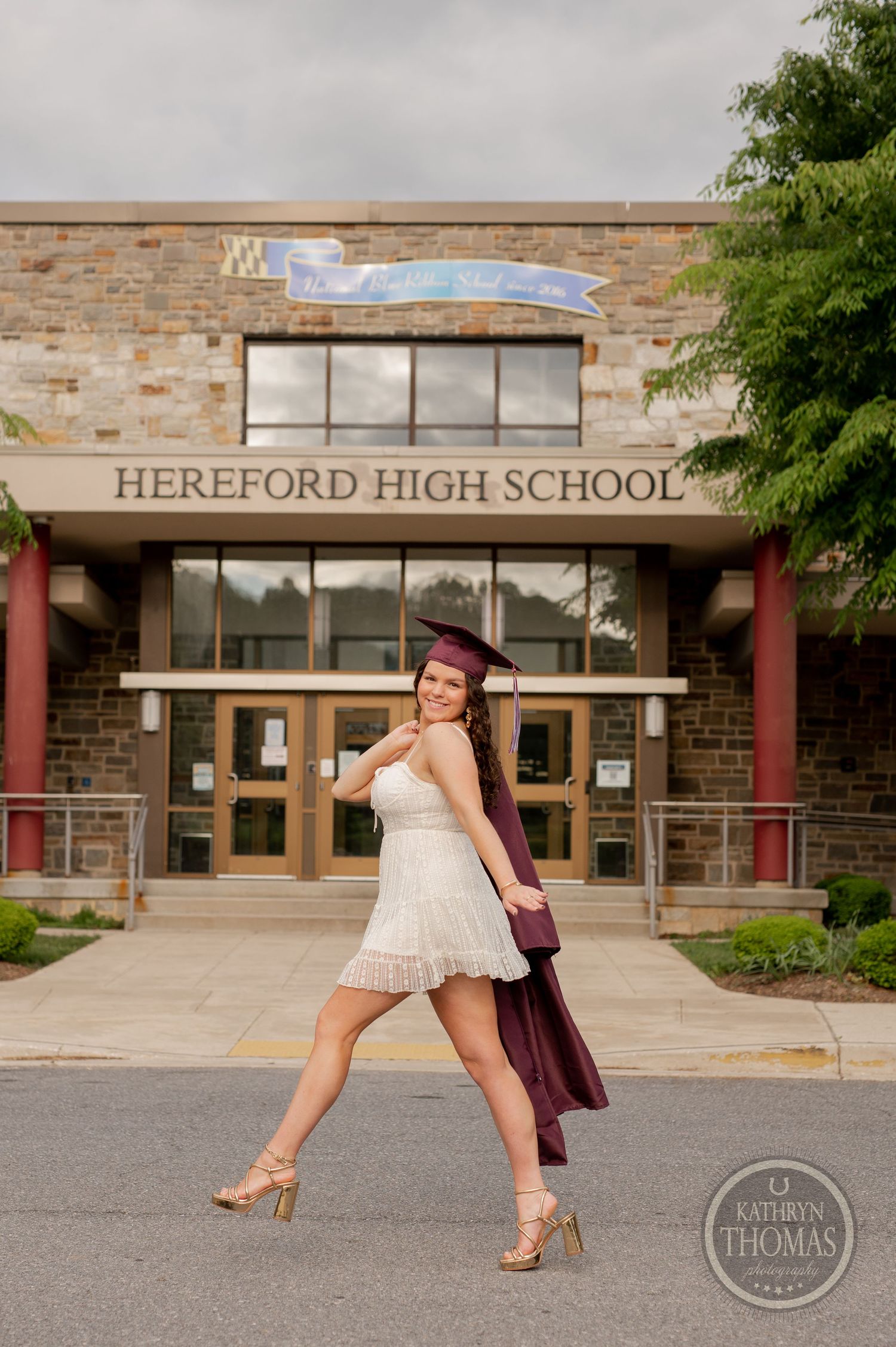 Graduate strikes playful pose in white dress and burgundy gown outside Hereford High School.