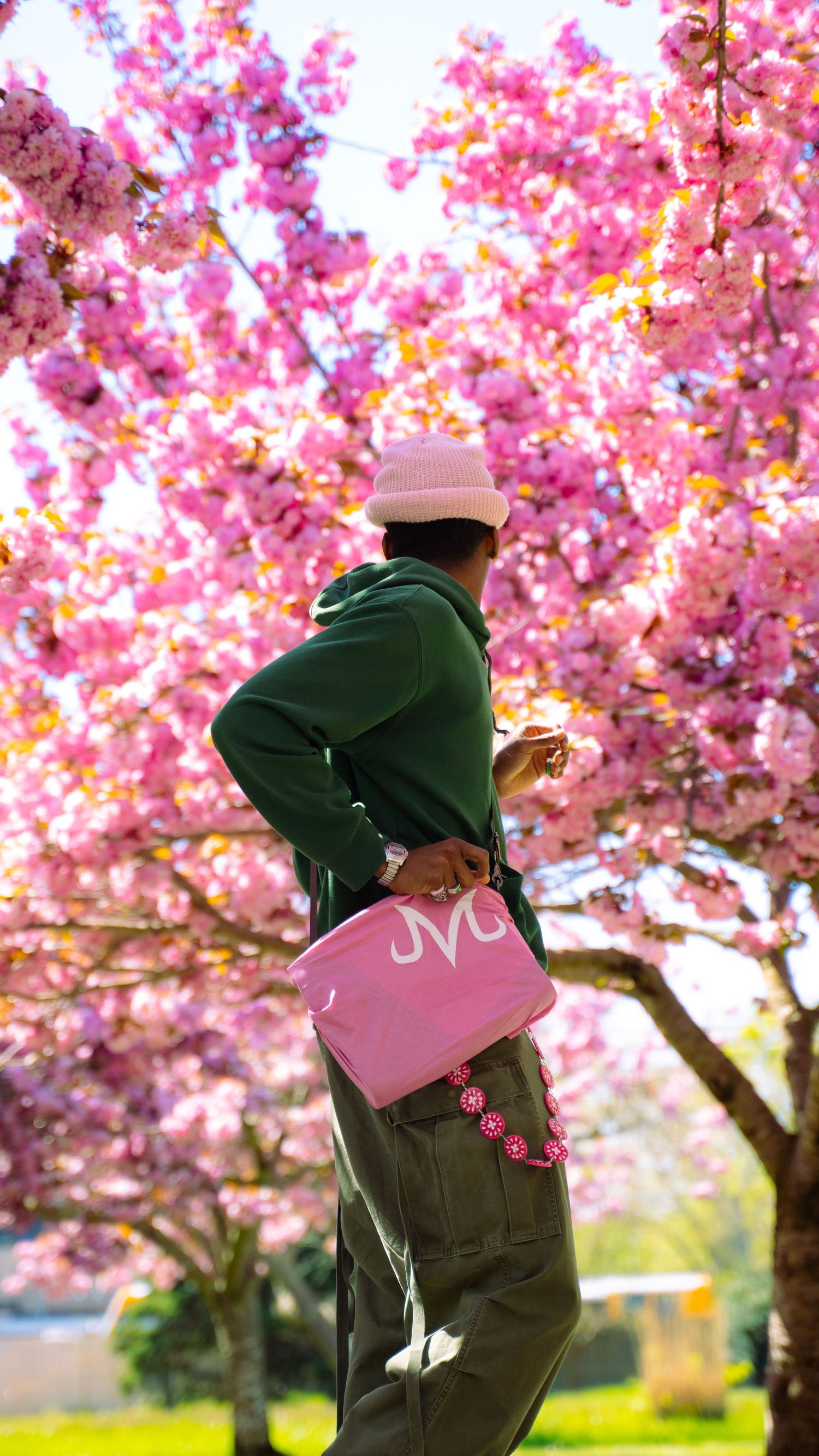 Person in green jacket and pink bag poses against vibrant cherry blossom trees in full bloom.