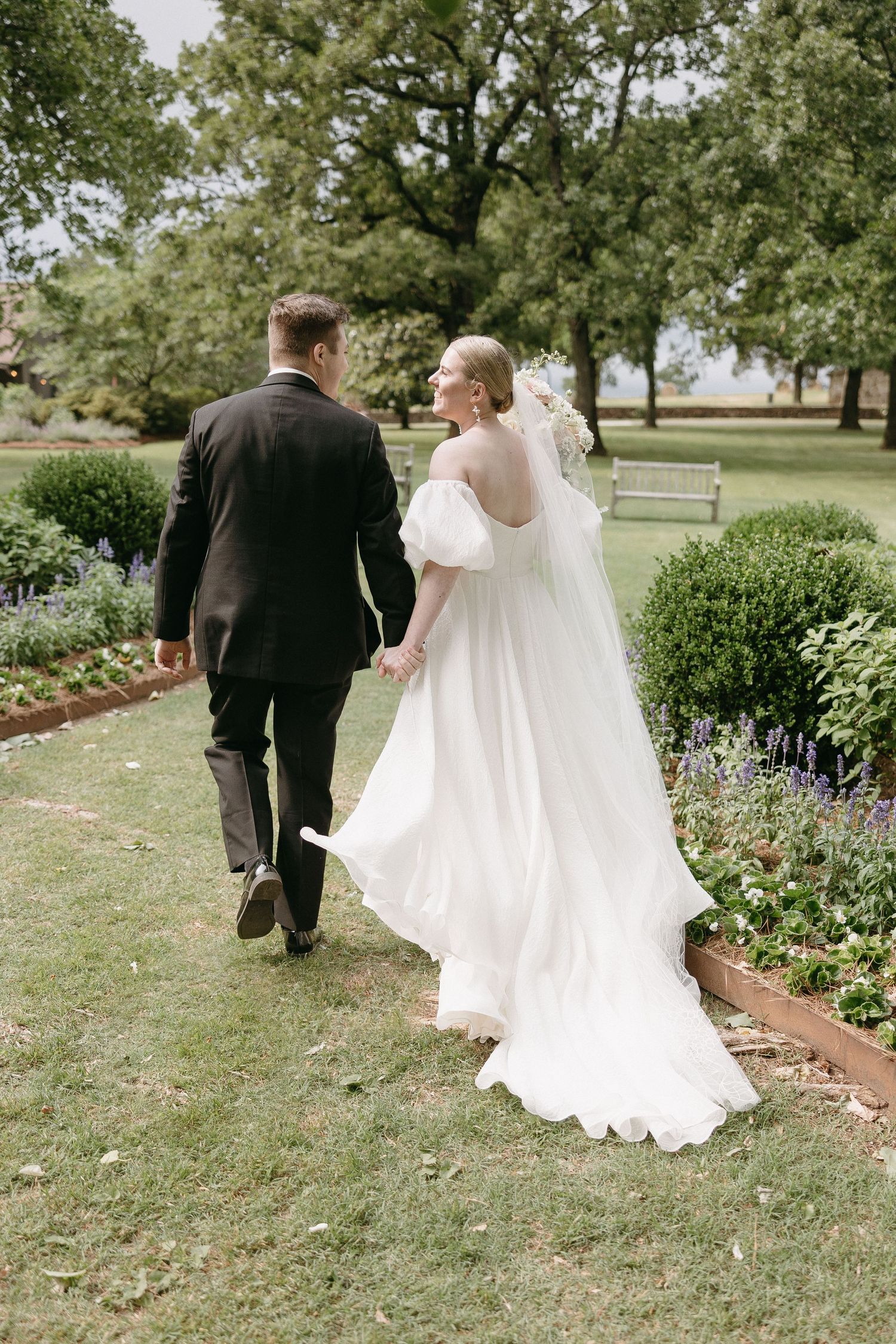 A couple walks hand in hand through a manicured garden path on their wedding day.