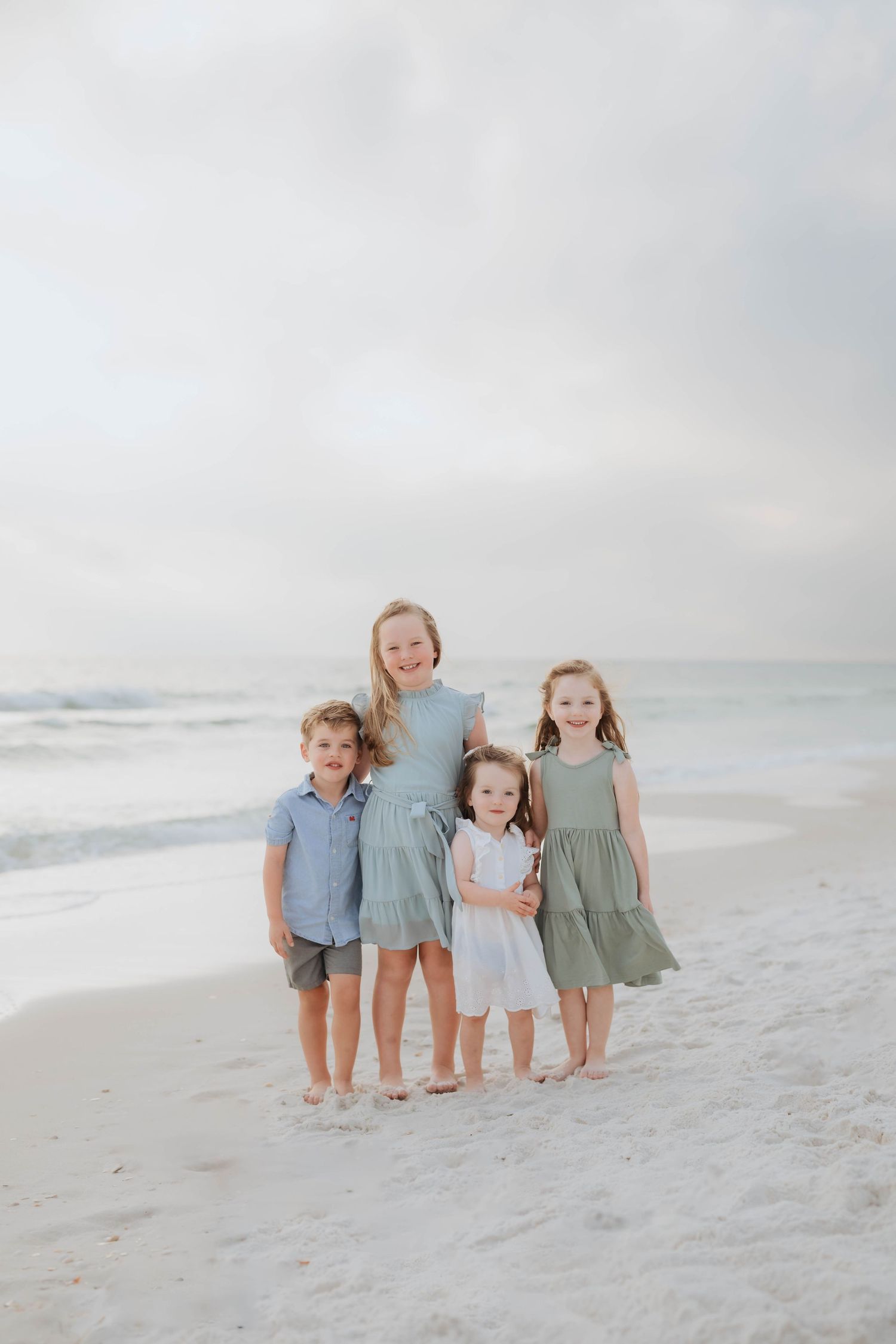 Four young children in soft pastel outfits stand together on a peaceful white sandy beach at sunset.