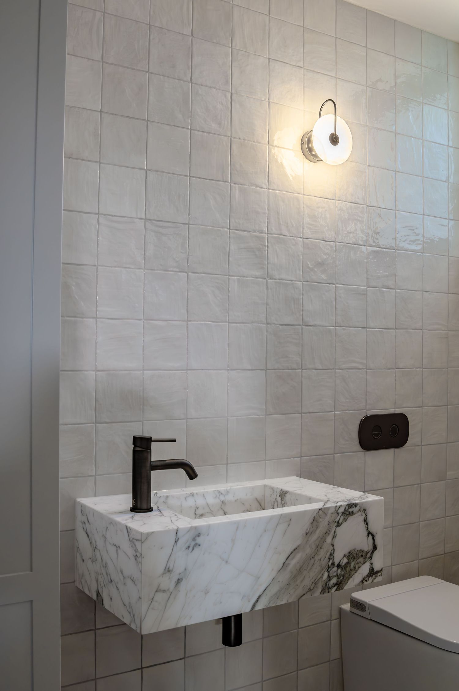 Modern powder room with marble floating sink, black fixtures, and white square tile walls with sconce lighting.