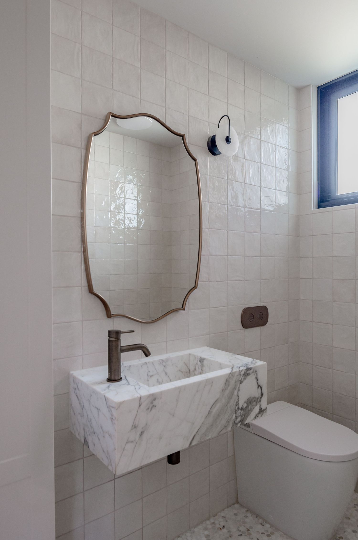 Powder room with floating marble sink, bronze-framed decorative mirror, and white square tile walls.
