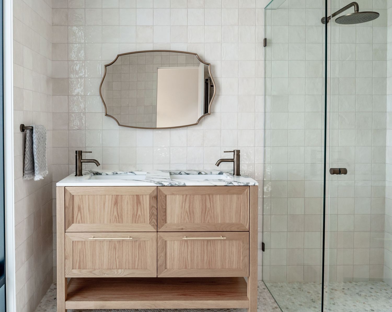 Modern bathroom with double wood vanity, frameless shower door, and decorative oval mirror.