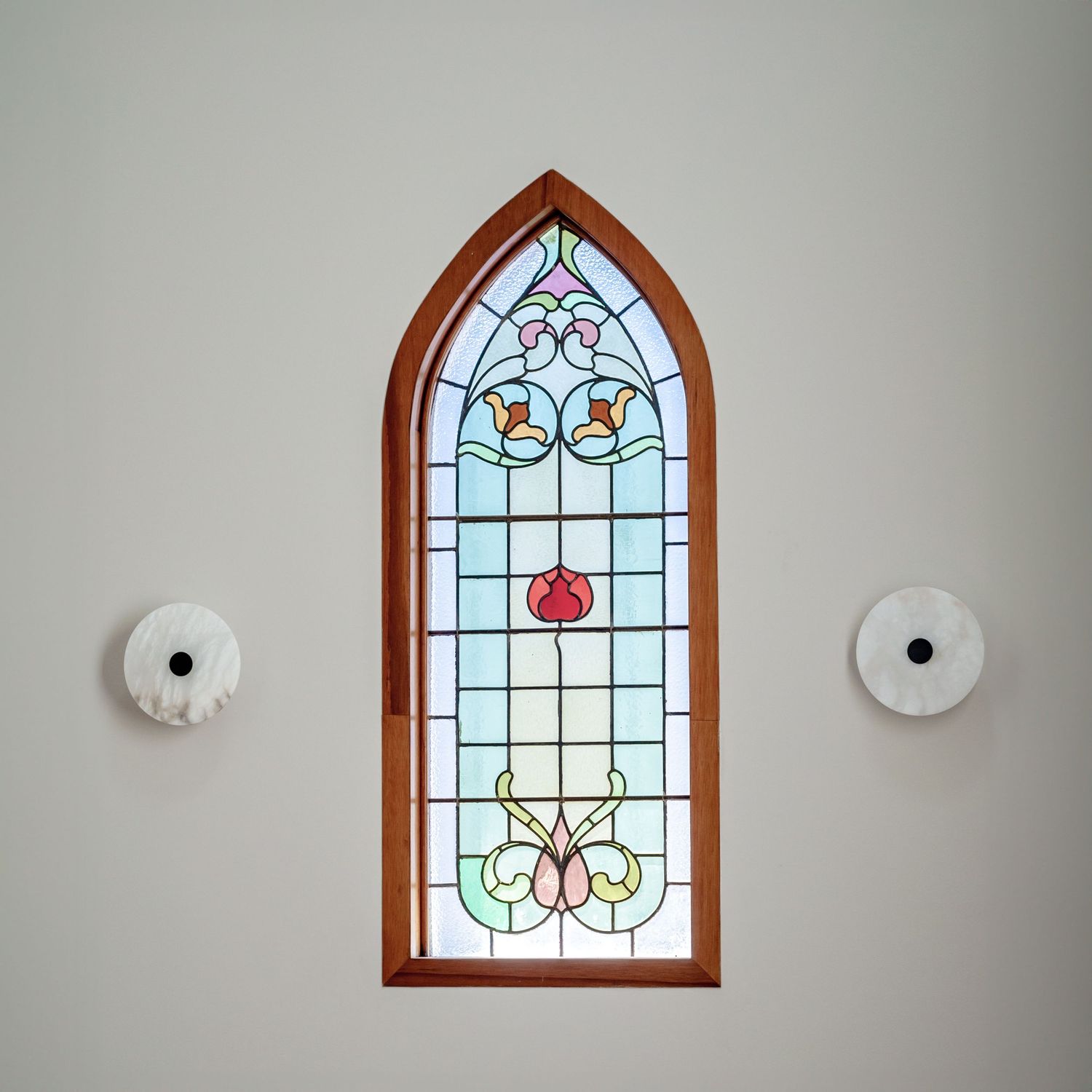 Elegant arched stained glass window with floral design flanked by two circular wall sconces.