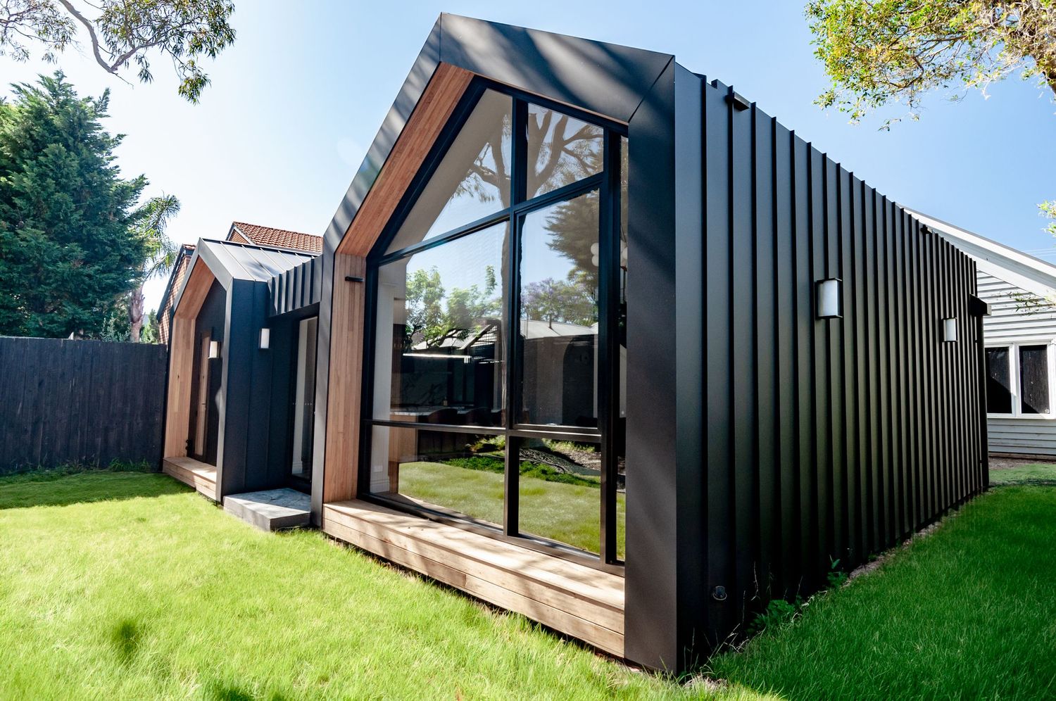 Modern black wooden garden studio with glass walls and wooden deck features an angled roof design in a backyard setting.