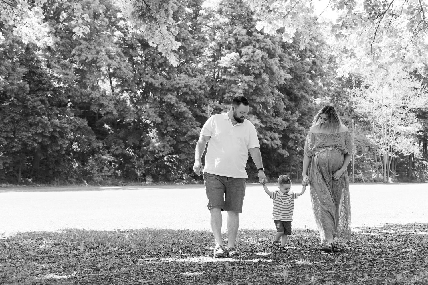 A family walks hand in hand along a wooded path on a sunny day captured in black and white photography.