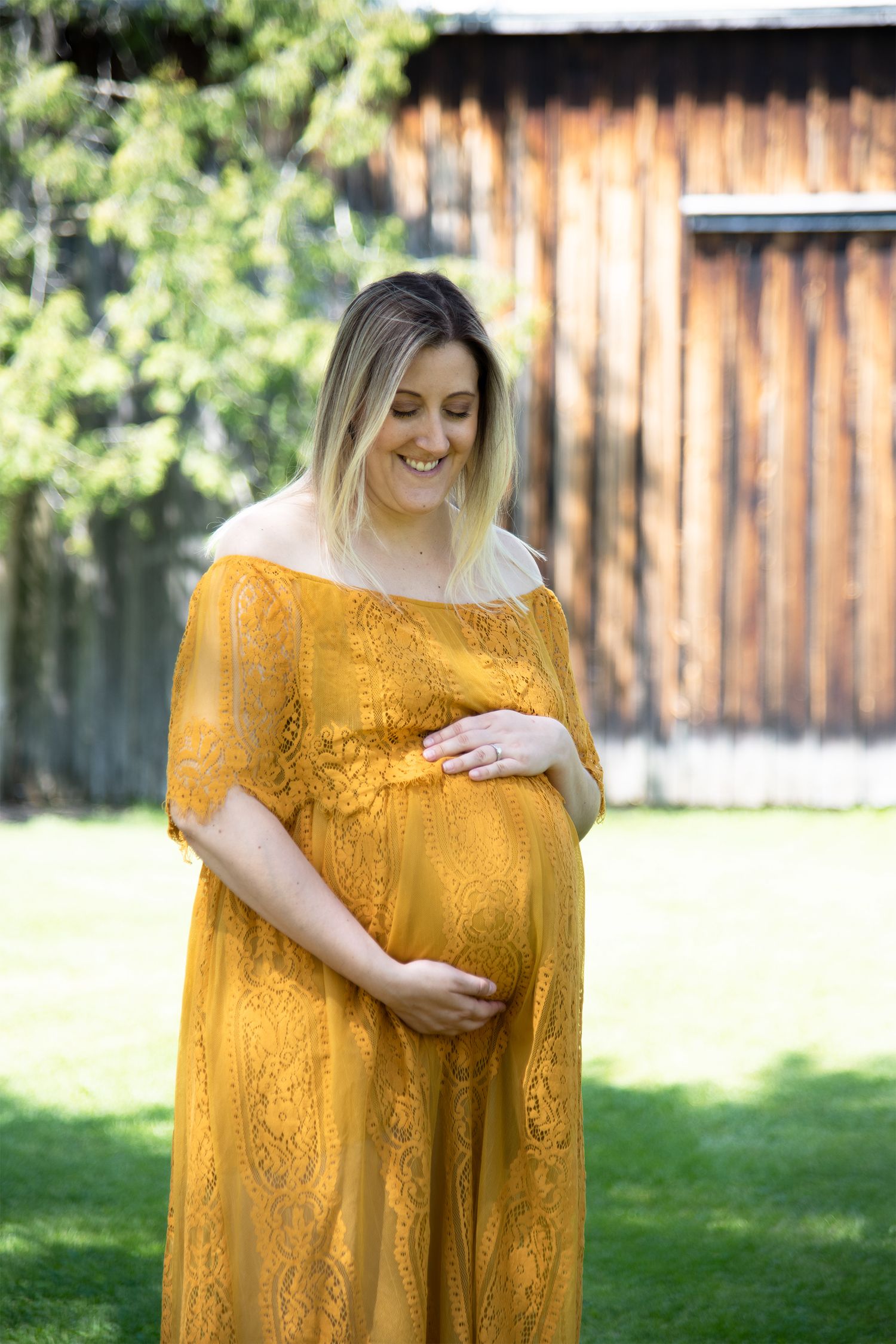 Woman in yellow lace dress cradles baby bump in outdoor portrait.