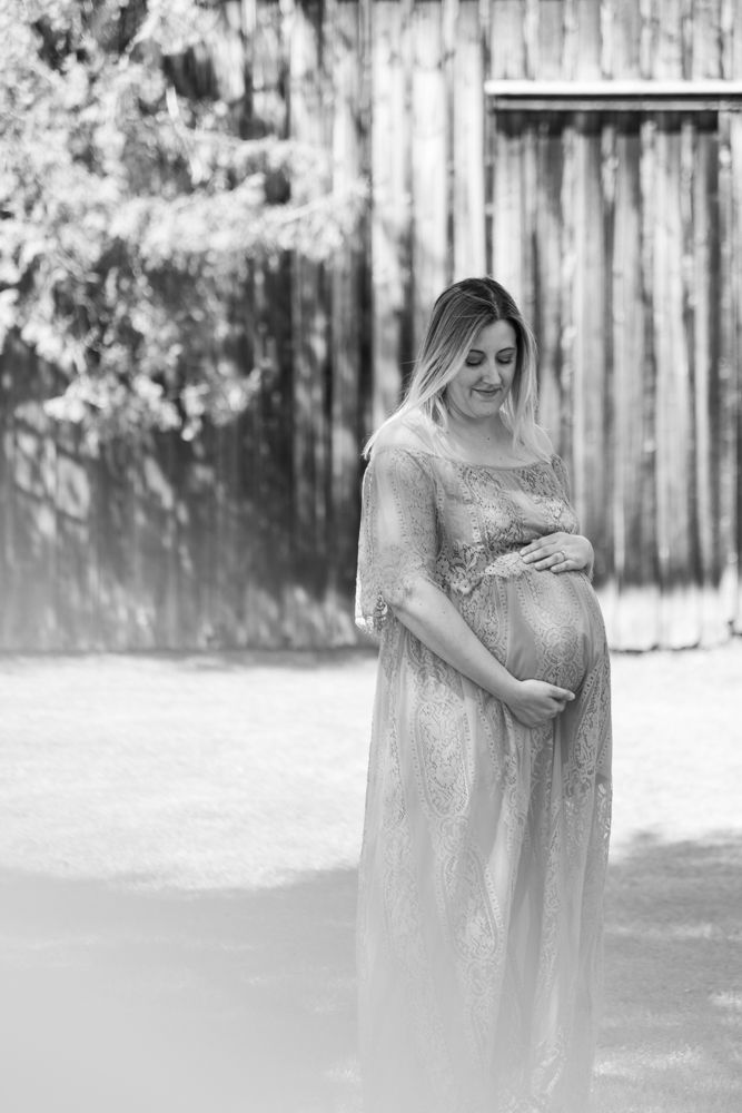 Black and white portrait of an expectant mother in a lace gown against a rustic wooden backdrop.