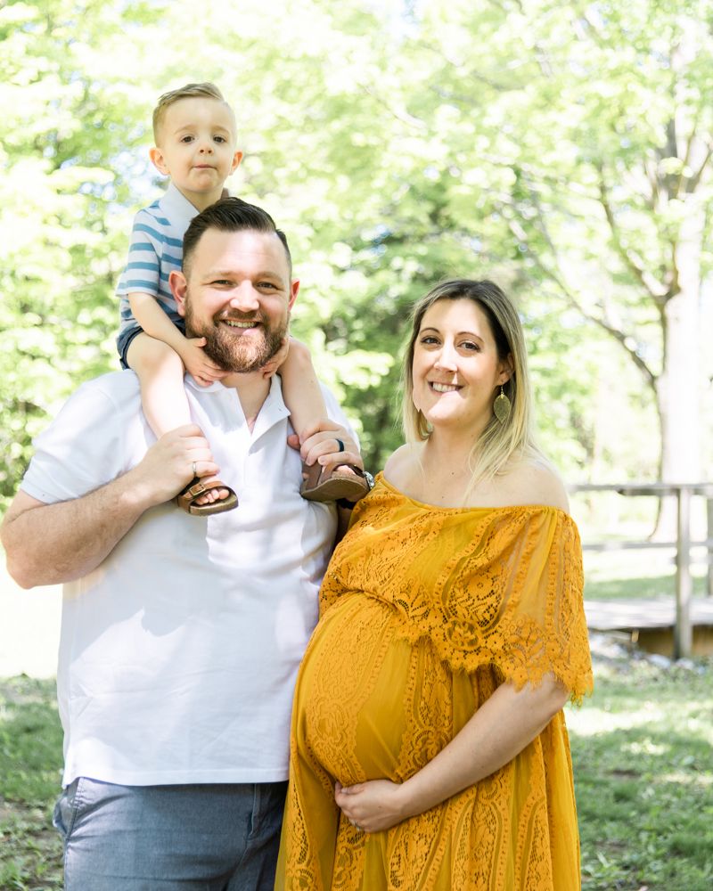 A couple poses for maternity photos, outdoors in a park setting, with a child perched on shoulders, and the woman wearing a yellow dress.