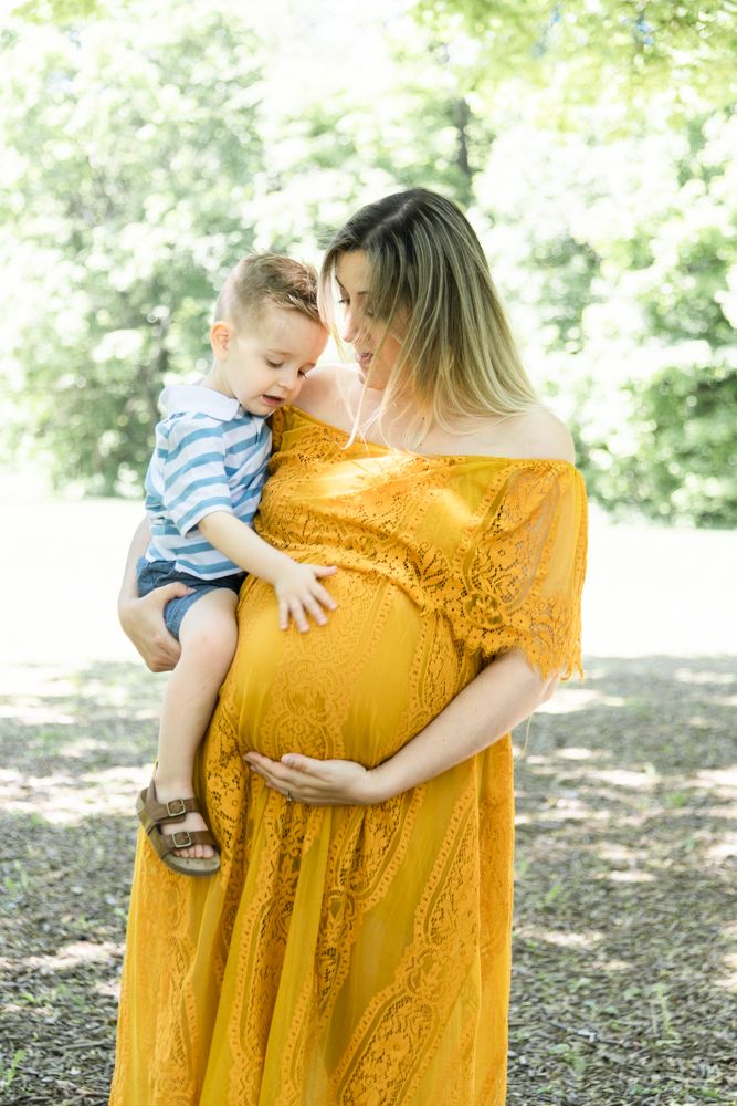 Mother in yellow lace dress holds toddler while displaying her baby bump in a sunlit outdoor setting.