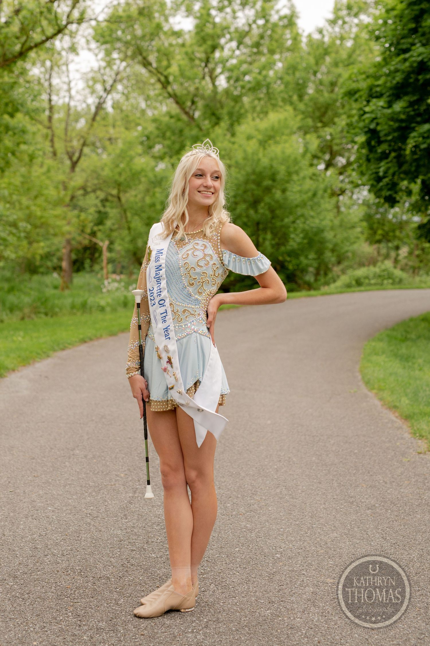 Beauty pageant contestant in sparkly dress and sash poses on walking path in park setting.