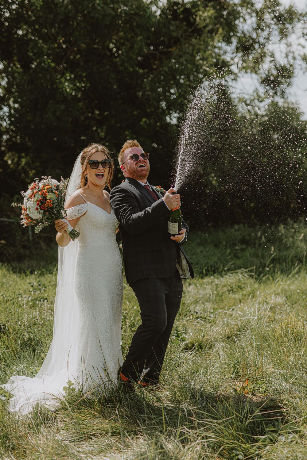 Newlyweds celebrating with champagne spray in a sunny outdoor field.