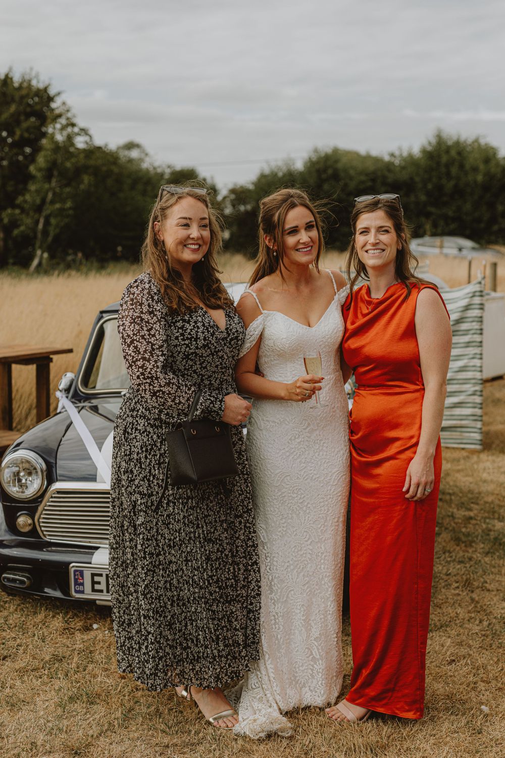 Three women in formal dresses pose together in front of a vintage car in a rustic outdoor setting.