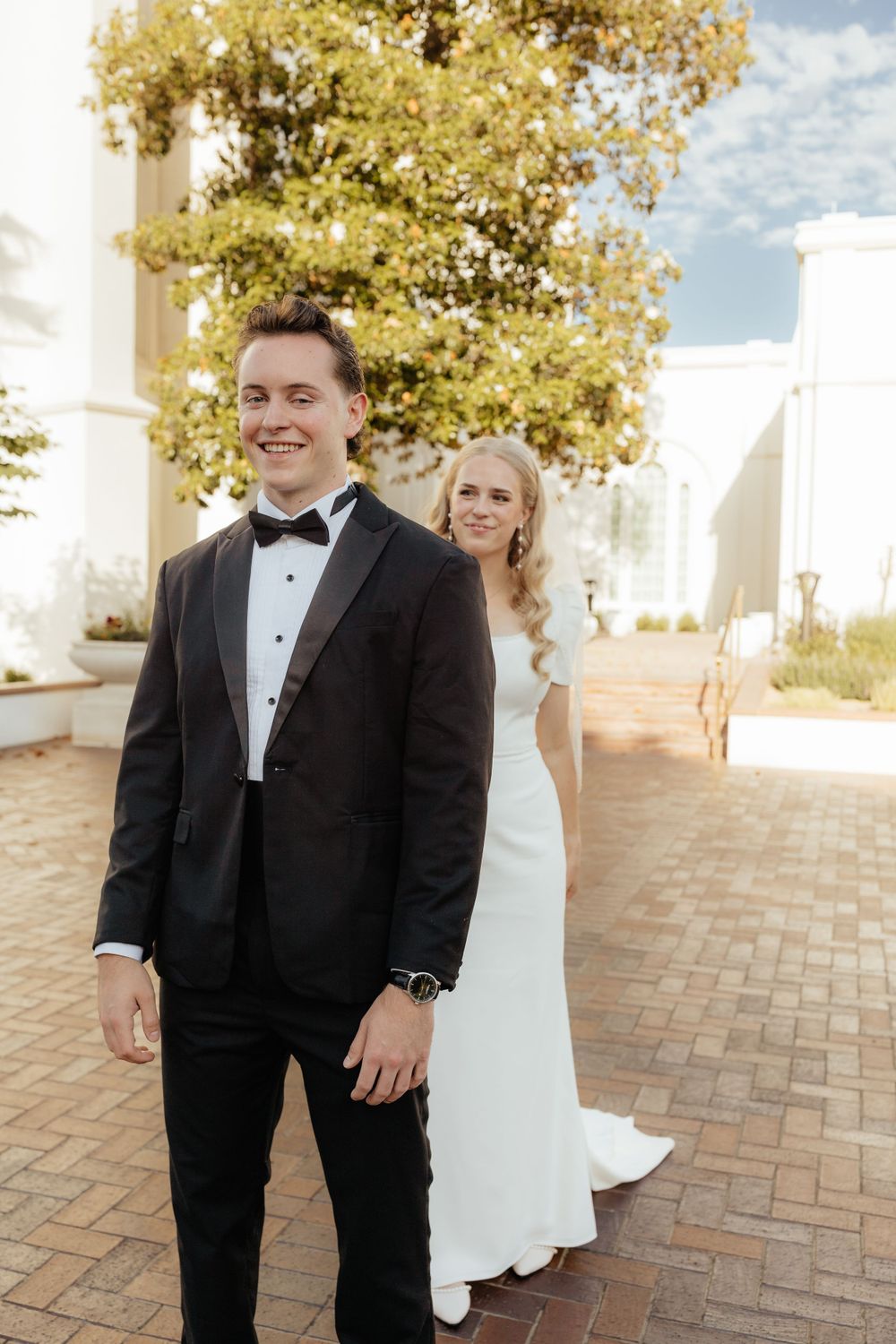 Smiling groom in tuxedo with bride in white dress posing outside white building.