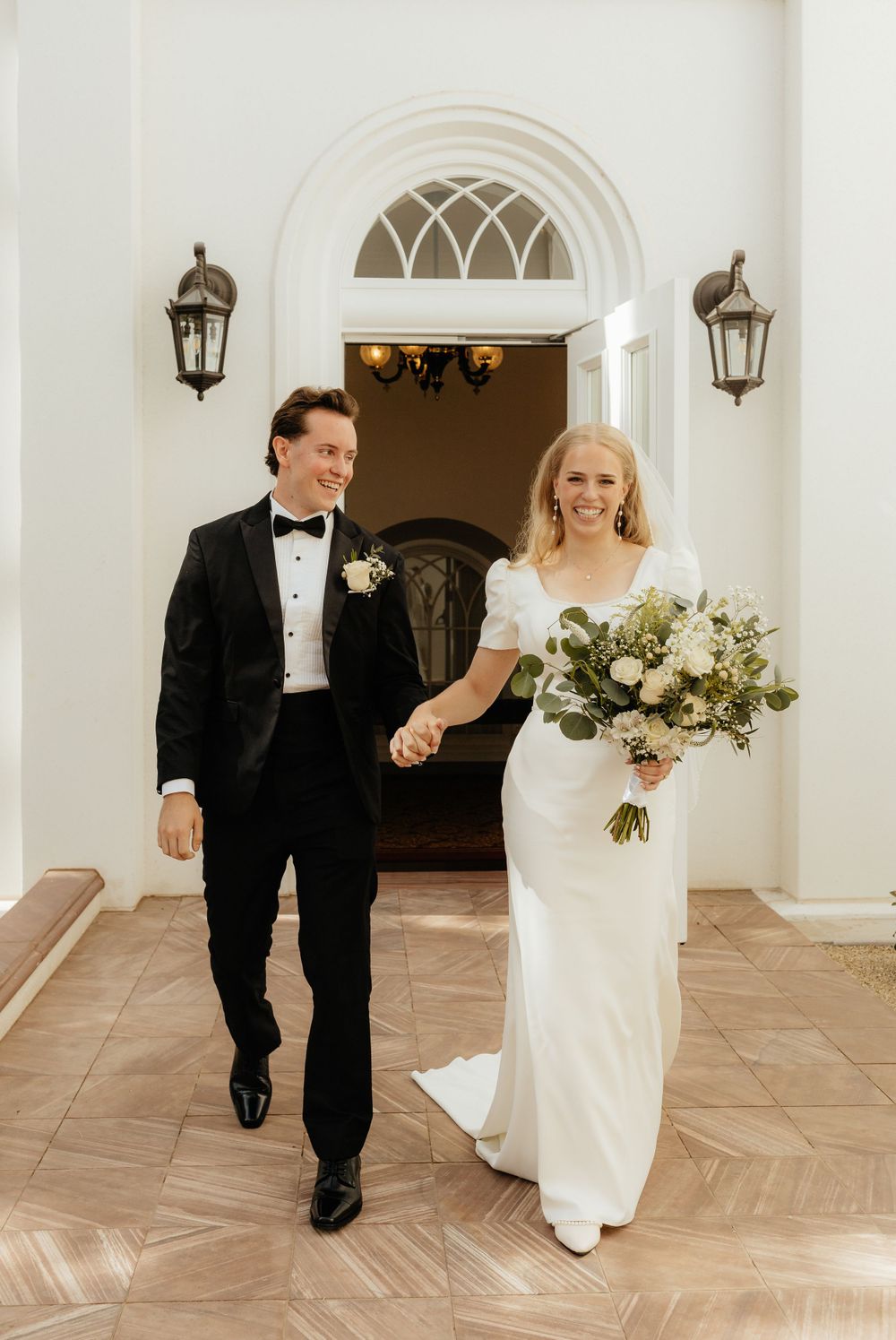 Elegant bride and groom walking hand in hand through grand arched doorway after wedding ceremony.