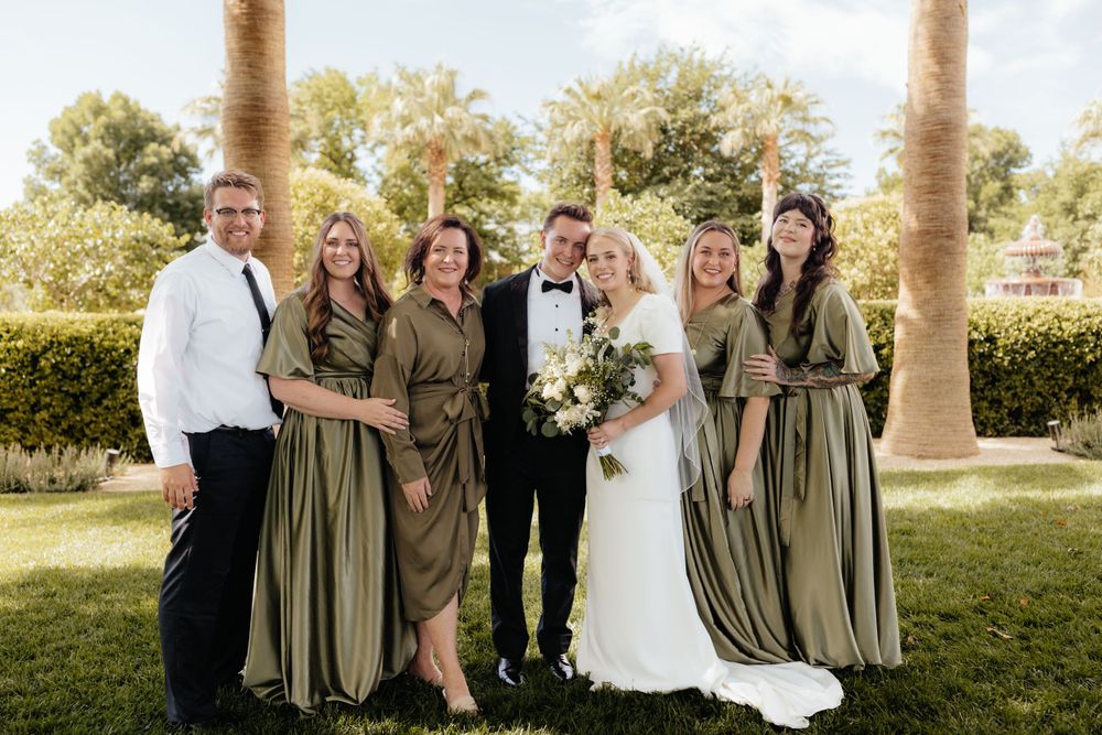 Bride and groom posing with bridesmaids in olive green dresses and groomsman outdoors near palm trees.