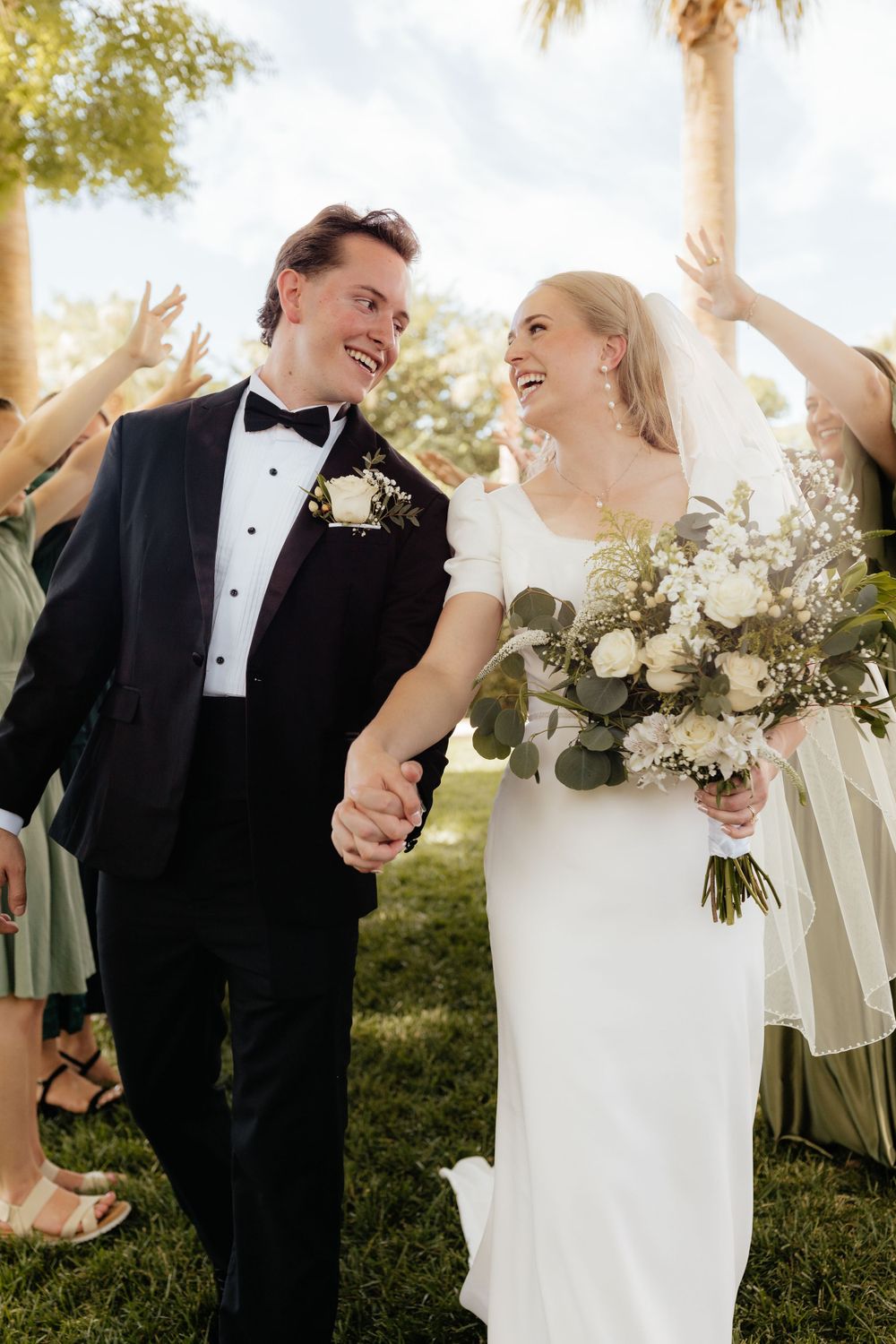 Joyful newlyweds holding hands walking through cheering guests outdoors with palm trees in background.