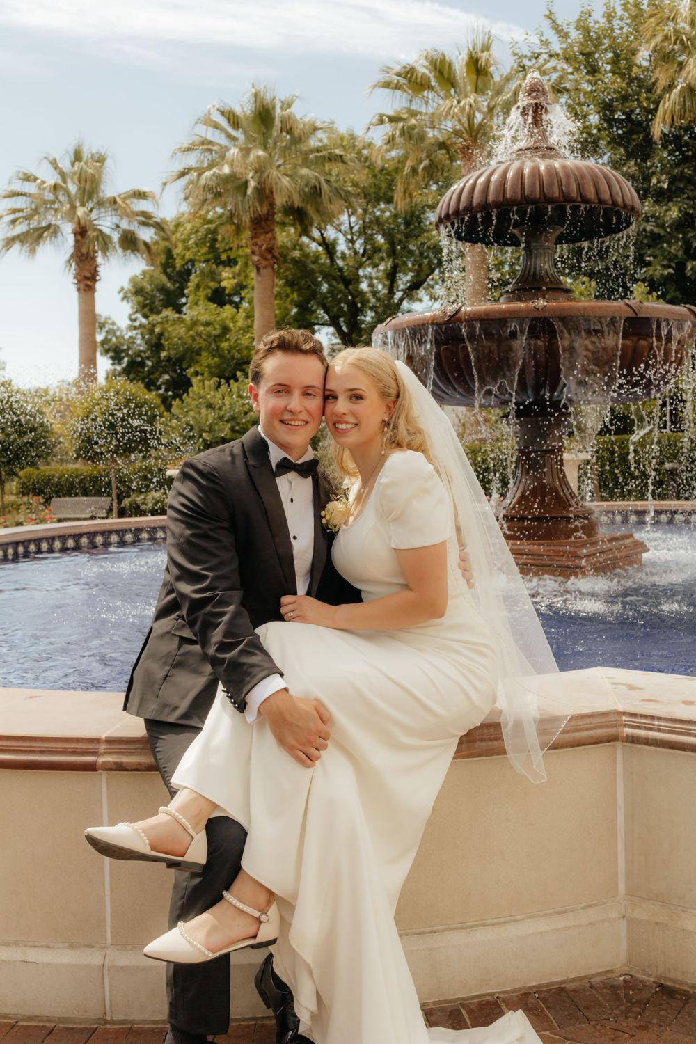 Smiling newlyweds posing together on fountain ledge in lush garden with palm trees and blue sky.