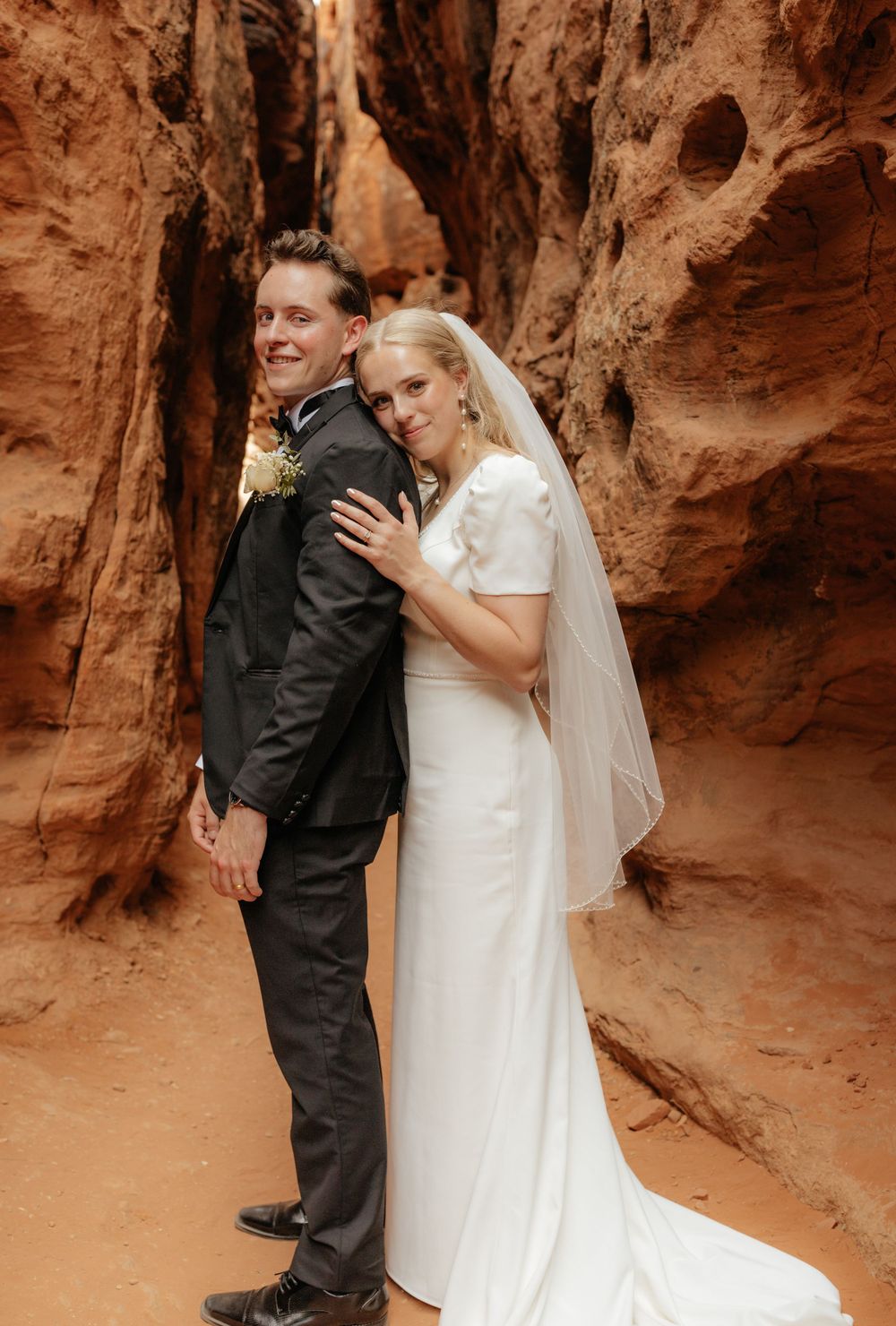Bride and groom posing back-to-back smiling in scenic red rock canyon setting.