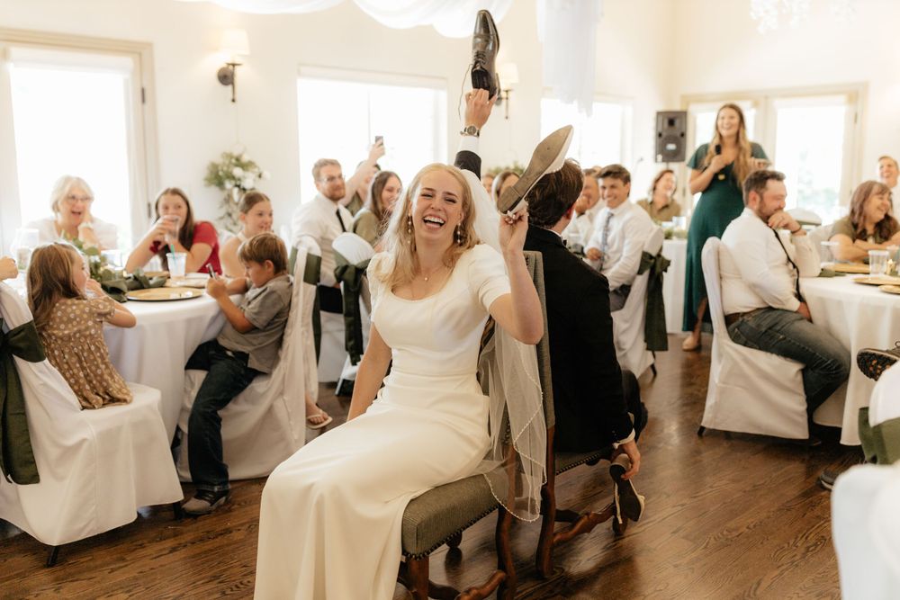 Laughing bride seated during garter toss game at lively wedding reception with guests cheering around her.