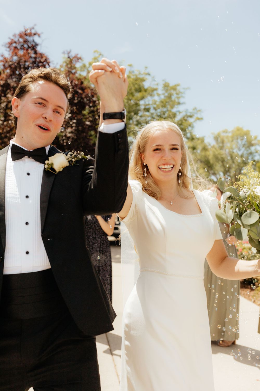 Joyful newlyweds raise hands in celebration after wedding ceremony outdoors on sunny day.