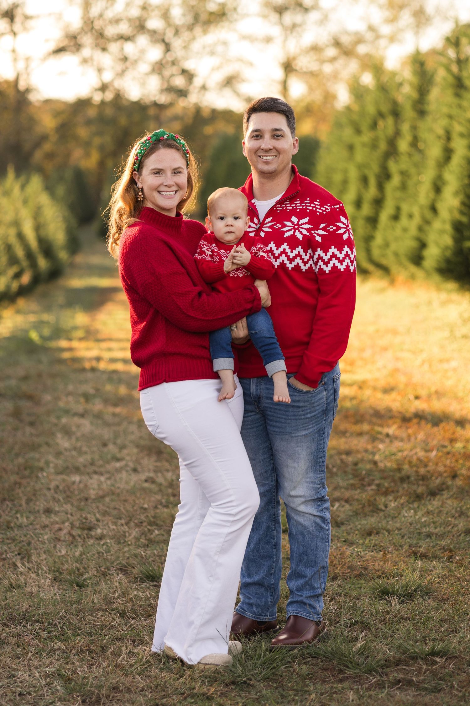 A family in matching red holiday sweaters poses together at a Christmas tree farm during golden hour.