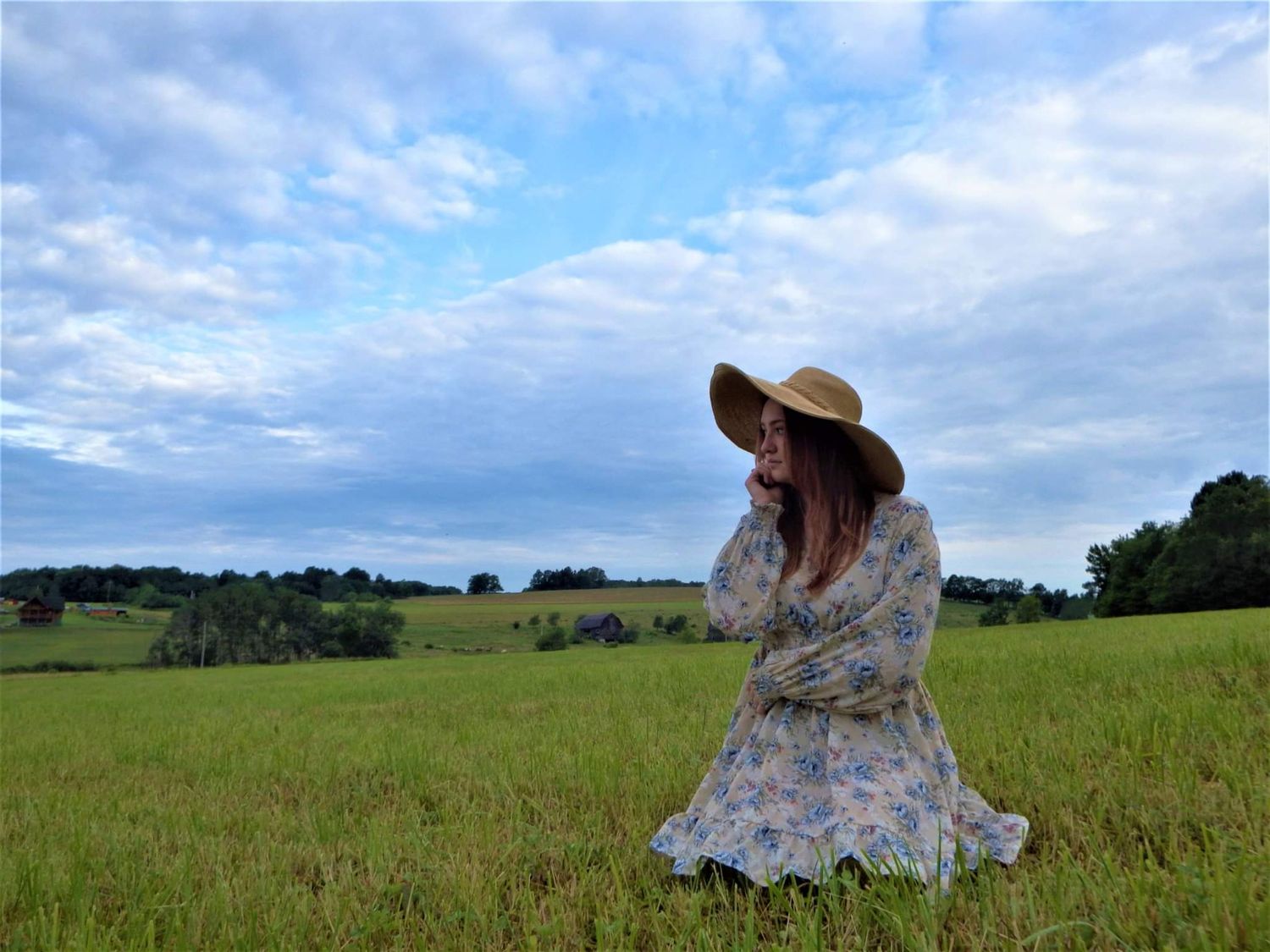 A person in a floral dress and sun hat sits peacefully in a green meadow under a cloudy summer sky.